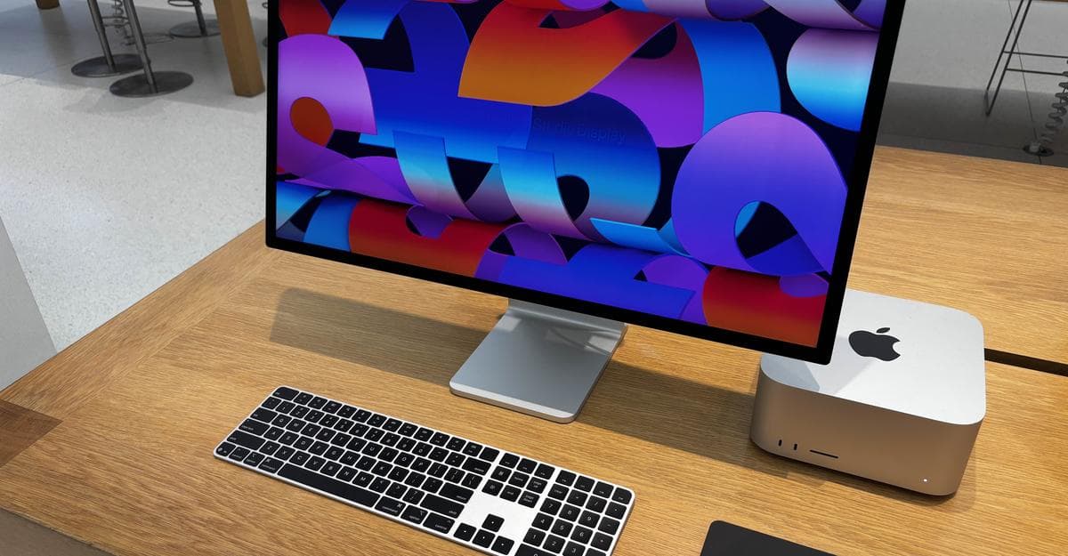 Apple Studio Display showing a colorful wallpaper beside a Mac Studio on a wooden desk, with a Magic Keyboard in front